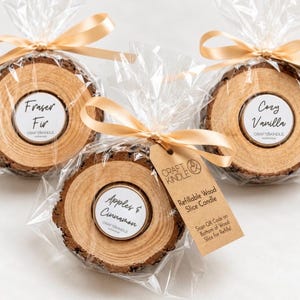 May include: Three refillable wood slice candles wrapped in clear plastic with gold ribbons. Labels read Fraser Fir, Cozy Vanilla, and Apples & Cinnamon. A brown tag features the text Craft Kindle and Refillable Wood Slice Candle.