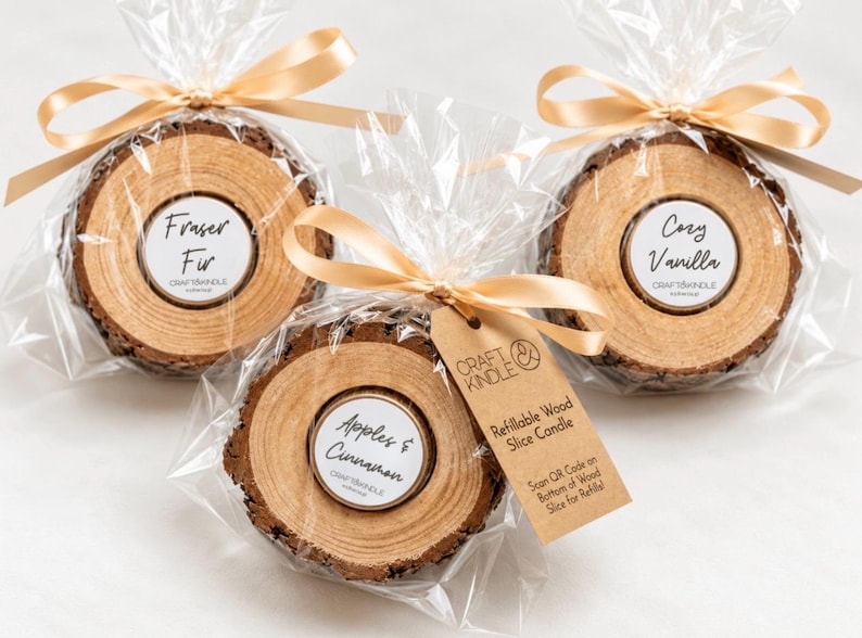 May include: Three refillable wood slice candles wrapped in clear plastic with gold ribbons. Labels read Fraser Fir, Cozy Vanilla, and Apples & Cinnamon. A brown tag features the text Craft Kindle and Refillable Wood Slice Candle.