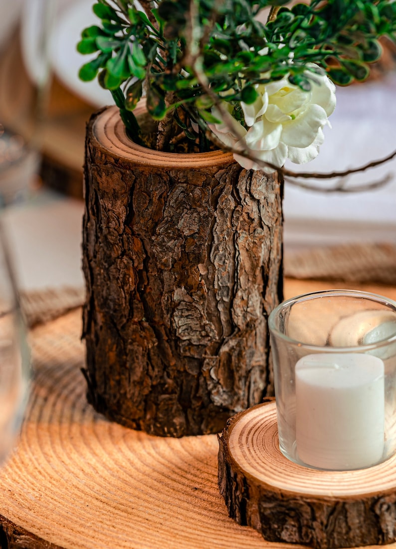 May include: A rustic wooden vase with a rough bark exterior holds greenery and a white flower. A small glass votive candle holder with a white candle sits on a wooden coaster, creating a natural, earthy aesthetic.