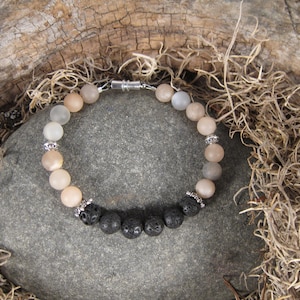 May include: A bracelet made with light brown, white, and gray gemstone beads and black lava rock beads. The bracelet has a silver clasp.