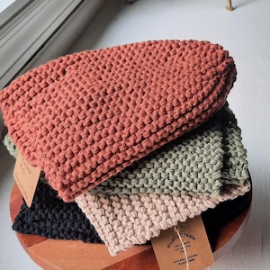 May include: Four knitted dishcloths stacked on a wooden stand. The dishcloths are in shades of brown, green, beige, and black. The top dishcloth has a brown tag with the text "Rustle & Needle" on it. The bottom dishcloth has a tag with the text "Rustle & Needle hand knit for you, enjoy your new dishcloths! www.rustleandneedle.com Rustle & Needle"