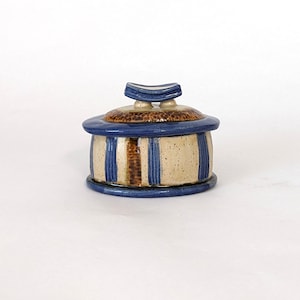 May include: A small, ceramic trinket box with a blue and brown striped design. The lid has a unique, curved shape and is also decorated with blue and brown accents.