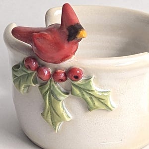 May include: A cream-colored ceramic yarn bowl featuring a sculpted red cardinal bird perched on a branch with green holly leaves and red berries. The bowl has a smooth, glossy finish and a U-shaped cutout on the rim for yarn to pass through.