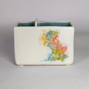 May include: A white ceramic utensil holder with a square shape and a small handle. The interior rim is a teal color. The front of the holder features a colorful abstract design with blue, yellow, orange, and pink hues.