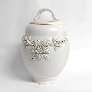 May include: White ceramic jar with a lid and a handle. The jar is decorated with raised floral designs.