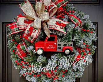 May include: A Christmas wreath with a red truck, plaid ribbons, and the words "Merry Christmas." The wreath is decorated with faux snow, red berries, and a large bow. The text "Notable Wreaths" is also visible.