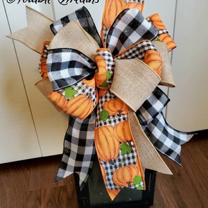 May include: A black lantern with a large bow made of burlap, black and white buffalo check, and orange pumpkin patterned ribbon. The bow is tied with a large loop and tails. The lantern is sitting on a wooden floor. "Notable Wreaths" is written in cursive on the top left of the image.
