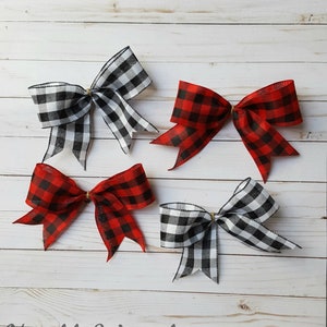 May include: Four decorative bows in a buffalo plaid pattern. Two bows are red and black, and two are black and white. The bows are tied with a gold-colored twine and are arranged on a white wooden surface. The text "Notable Wreaths" is at the bottom.