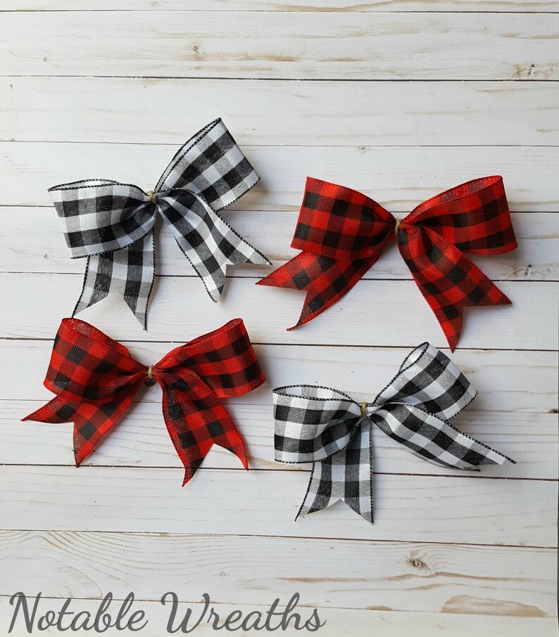 May include: Four decorative bows in a buffalo plaid pattern. Two bows are red and black, and two are black and white. The bows are tied with a gold-colored wire and are arranged on a white wooden surface. The text "Notable Wreaths" is at the bottom.