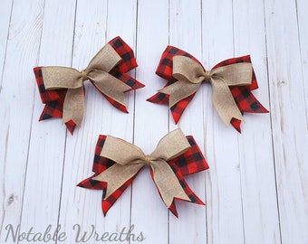 May include: Three decorative bows with a red and black buffalo plaid pattern and a tan burlap bow. The bows are arranged on a white wooden surface. The text "Notable Wreaths" is at the bottom.