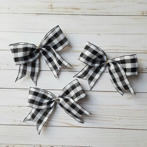 May include: Three decorative bows with a black and white checkered pattern. The bows are tied with a thin, light-colored twine and have a black trim. The bows are arranged on a light-colored wooden surface. The text "Notable Wreaths" is at the bottom.