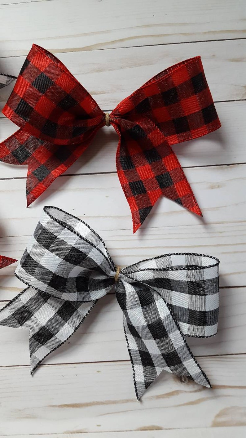 May include: Two decorative bows, one red and black buffalo plaid, the other black and white. The bows are tied with twine and have a classic, rustic aesthetic. The fabric has a woven texture.