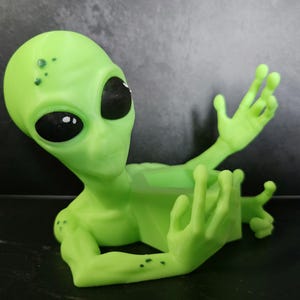 Puede incluir: Una figura de alien&iacute;gena verde claro con grandes ojos negros y brazos extendidos. El alien&iacute;gena tiene peque&ntilde;as manchas oscuras en la cabeza y los brazos. La figura est&aacute; hecha de un material liso y descansa sobre una superficie oscura.