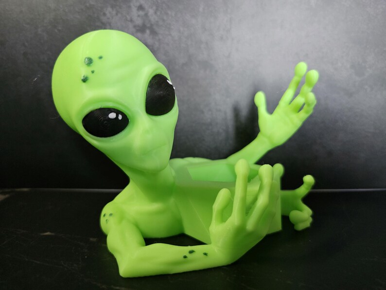 Puede incluir: Una figura de alien&iacute;gena verde claro con grandes ojos negros y brazos extendidos. El alien&iacute;gena tiene peque&ntilde;as manchas oscuras en la cabeza y los brazos. La figura est&aacute; hecha de un material liso y descansa sobre una superficie oscura.