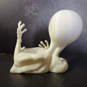 Puede incluir: Una maceta con forma de alien&iacute;gena de color crema. La maceta tiene una cabeza grande, brazos extendidos y una abertura rectangular para plantas. El alien&iacute;gena est&aacute; acostado, con la cabeza y los brazos levantados.