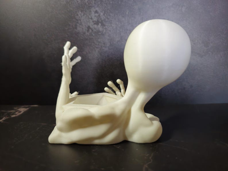 Puede incluir: Una maceta con forma de alien&iacute;gena de color crema. La maceta tiene una cabeza grande, brazos extendidos y una abertura rectangular para plantas. El alien&iacute;gena est&aacute; acostado, con la cabeza y los brazos levantados.