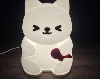 Puede incluir: Una luz de noche blanca con forma de gato con un detalle rojo en forma de corazón. La lámpara tiene un diseño lindo con orejas triangulares, una nariz pequeña y ojos. La lámpara se ilumina desde adentro, creando un brillo suave.