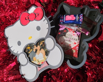 Puede incluir: Un contenedor gris y blanco con forma de Hello Kitty con un lazo rojo, que contiene una foto y caramelos. La foto es de una pareja. El contenedor está lleno de caramelos envueltos, incluyendo una galleta en forma de corazón.