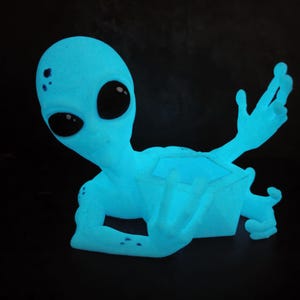 Puede incluir: Figura de alien&iacute;gena azul brillante con grandes ojos negros y brazos extendidos. El alien&iacute;gena tiene una textura suave y est&aacute; sobre un fondo negro. La figura parece ser un objeto decorativo.