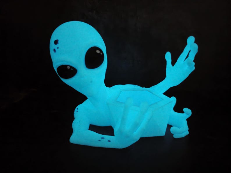 Puede incluir: Figura de alien&iacute;gena azul brillante con grandes ojos negros y brazos extendidos. El alien&iacute;gena tiene una textura suave y est&aacute; sobre un fondo negro. La figura parece ser un objeto decorativo.