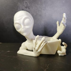 Puede incluir: Una maceta con forma de alien&iacute;gena de color crema, con ojos grandes y brazos extendidos. La maceta tiene un recipiente geom&eacute;trico abierto para guardar peque&ntilde;as plantas u objetos decorativos. La figura alien&iacute;gena est&aacute; posada como si emergiera del recipiente.