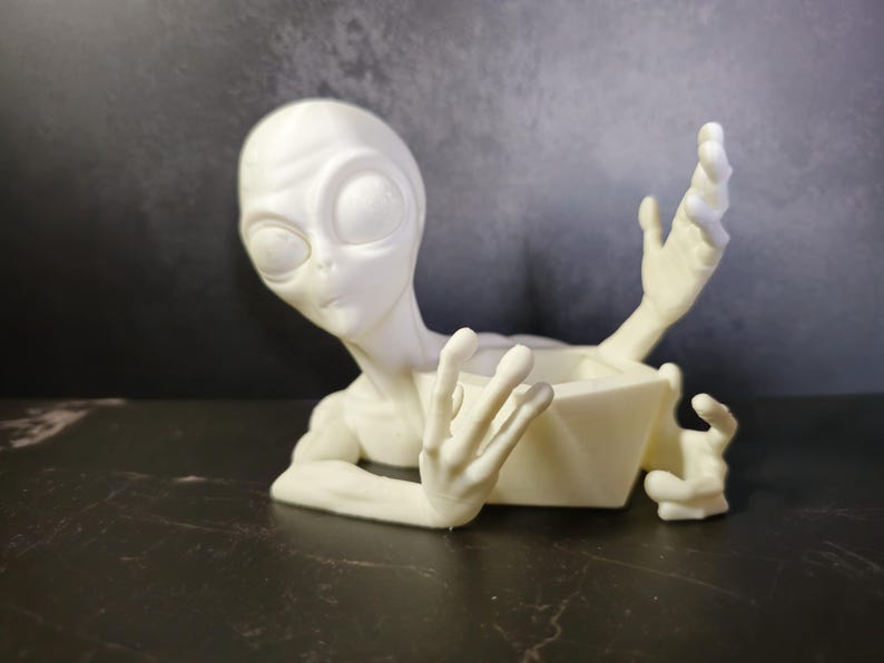 Puede incluir: Una maceta con forma de alien&iacute;gena de color crema, con ojos grandes y brazos extendidos. La maceta tiene un recipiente geom&eacute;trico abierto para guardar peque&ntilde;as plantas u objetos decorativos. La figura alien&iacute;gena est&aacute; posada como si emergiera del recipiente.