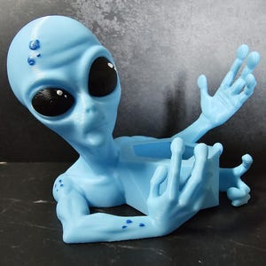 Puede incluir: Figura de alien&iacute;gena azul claro con grandes ojos negros y brazos extendidos. El alien&iacute;gena tiene una textura suave y sostiene un peque&ntilde;o recipiente rectangular. Un art&iacute;culo decorativo &uacute;nico.