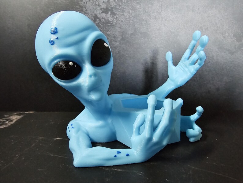 Puede incluir: Figura de alien&iacute;gena azul claro con grandes ojos negros y brazos extendidos. El alien&iacute;gena tiene una textura suave y sostiene un peque&ntilde;o recipiente rectangular. Un art&iacute;culo decorativo &uacute;nico.