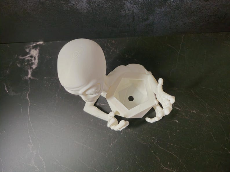Puede incluir: Una maceta blanca con forma de alien&iacute;gena impresa en 3D. La maceta presenta una cabeza grande, ojos peque&ntilde;os y brazos y manos extendidos. El cuerpo de la maceta tiene una forma geom&eacute;trica con un agujero central para plantar. El dise&ntilde;o es caprichoso y &uacute;nico.