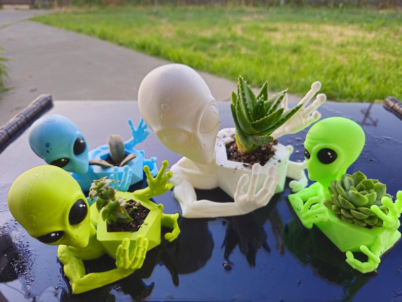 Puede incluir: Una colecci&oacute;n de macetas con forma de alien&iacute;gena en varios colores, incluyendo azul, verde y blanco. Cada maceta contiene una peque&ntilde;a planta suculenta o aloe vera. Las macetas est&aacute;n dispuestas sobre una superficie oscura, con un fondo de hierba.