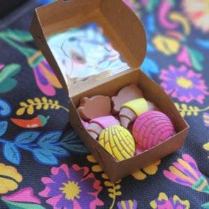 May include: A brown cardboard box with a clear lid contains several colorful pastries. The pastries include chocolate-colored croissants with yellow and pink accents, and round, pink and yellow concha bread. The box sits on a floral patterned fabric.