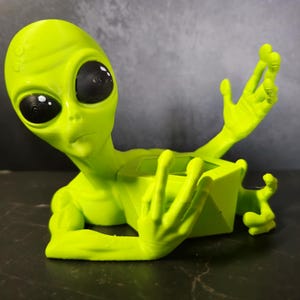 Puede incluir: Una figura de alien&iacute;gena verde lima con grandes ojos negros y brazos extendidos. El alien&iacute;gena est&aacute; en posici&oacute;n reclinada, con un recipiente cuadrado en su secci&oacute;n media. La figura est&aacute; hecha de un material liso.
