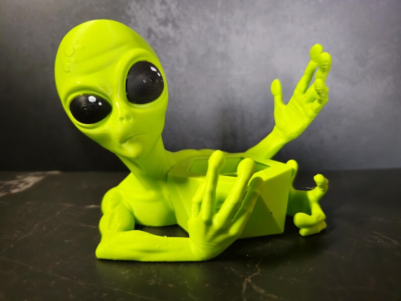 Puede incluir: Una figura de alien&iacute;gena verde lima con grandes ojos negros y brazos extendidos. El alien&iacute;gena est&aacute; en posici&oacute;n reclinada, con un recipiente cuadrado en su secci&oacute;n media. La figura est&aacute; hecha de un material liso.