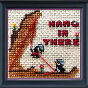 May include: Cross-stitch embroidery with two penguins climbing a cliff with red ropes. The text "HANG IN THERE" is stitched in red on a white background.