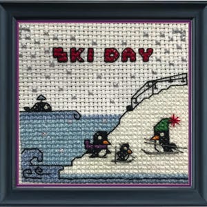 May include: A cross-stitch design featuring a snowy scene with penguins skiing down a hill. The text "SKI DAY" is stitched in red and white letters at the top of the design.