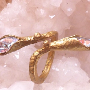 Puede incluir: Un anillo de oro con dos piedras de cristal iridiscentes engastadas en los extremos del anillo. El anillo está hecho de metal texturizado y tiene una forma orgánica única.