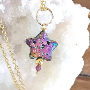May include: A gold chain necklace with a star-shaped pendant made of iridescent purple and black stone. The pendant is accented with a gold bead and a small pink gemstone.