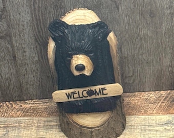 May include: A carved wooden bear head with a black painted face and a wooden sign that says "WELCOME" with a paw print.