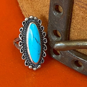 Puede incluir: Un anillo de plata con una piedra turquesa engastada en forma ovalada. El anillo est&aacute; rodeado por un borde decorativo de plata.