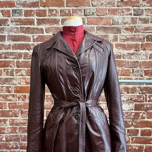May include: A brown leather jacket with a belt, a button closure, and a pointed collar. The jacket has a fitted silhouette and is lined with a brown fabric.