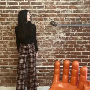 May include: A woman wearing a black turtleneck sweater and brown and white plaid pants stands in front of a brick wall. A large orange hand-shaped chair is in the foreground.
