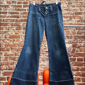 Pu&ograve; includere: Un paio di jeans a zampa di elefante in denim blu scuro con chiusura a bottoni e grandi tasche. I jeans hanno un'apertura della gamba larga e sono appesi a un appendiabiti.