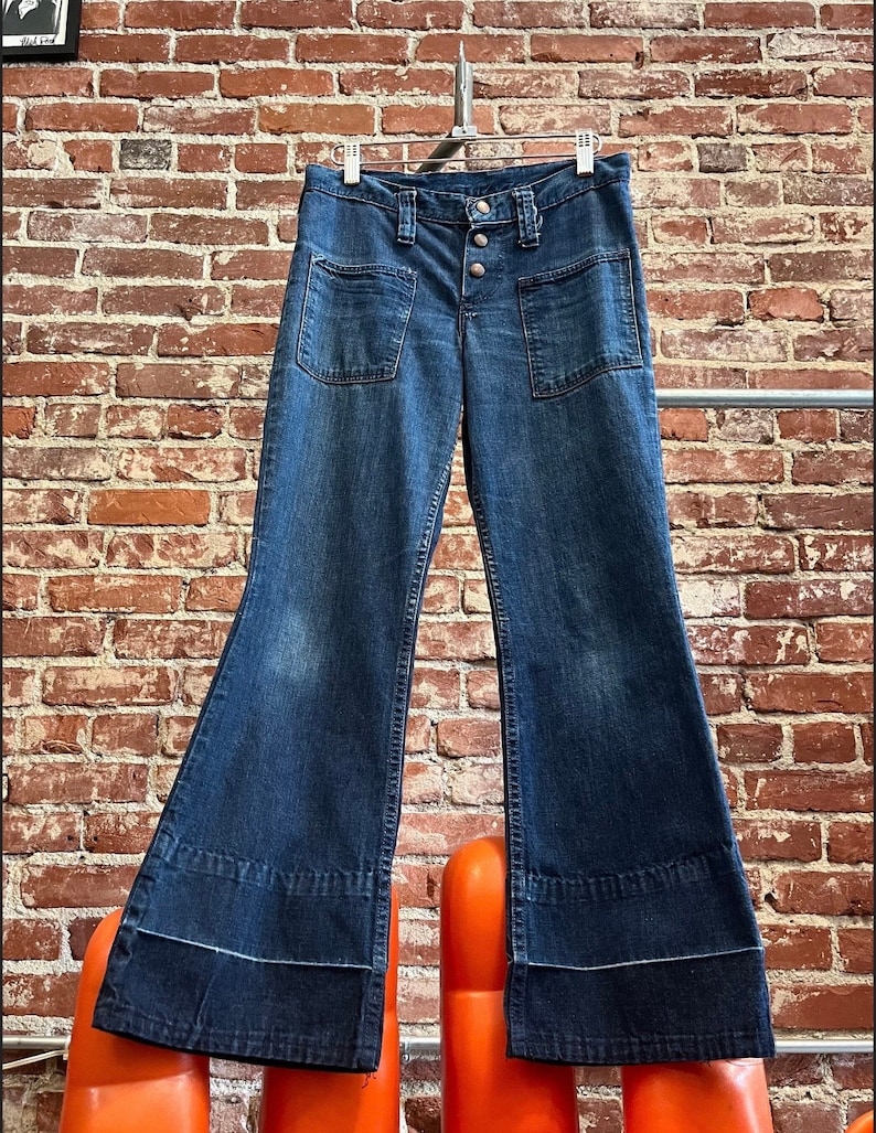 Pu&ograve; includere: Un paio di jeans a zampa di elefante in denim blu scuro con chiusura a bottoni e grandi tasche. I jeans hanno un'apertura della gamba larga e sono appesi a un appendiabiti.