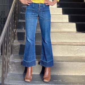 Pu&ograve; includere: Un paio di jeans a campana blu in denim con chiusura a bottone e due grandi tasche su ogni gamba. I jeans sono indossati da una persona in piedi su una rampa di scale.