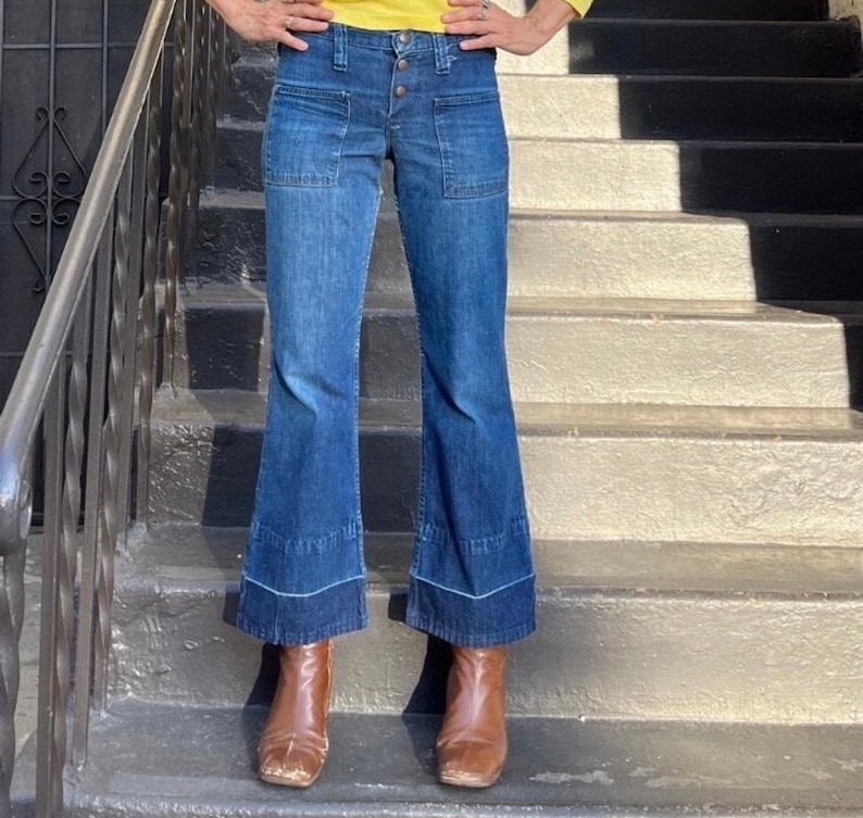 Pu&ograve; includere: Un paio di jeans a campana blu in denim con chiusura a bottone e due grandi tasche su ogni gamba. I jeans sono indossati da una persona in piedi su una rampa di scale.