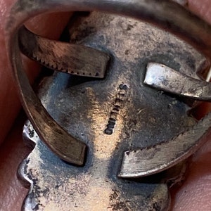 Puede incluir: Primer plano de la parte posterior de un anillo de plata con una inscripci&oacute;n estampada que dice "STERLING". El anillo tiene un dise&ntilde;o detallado con un borde festoneado.