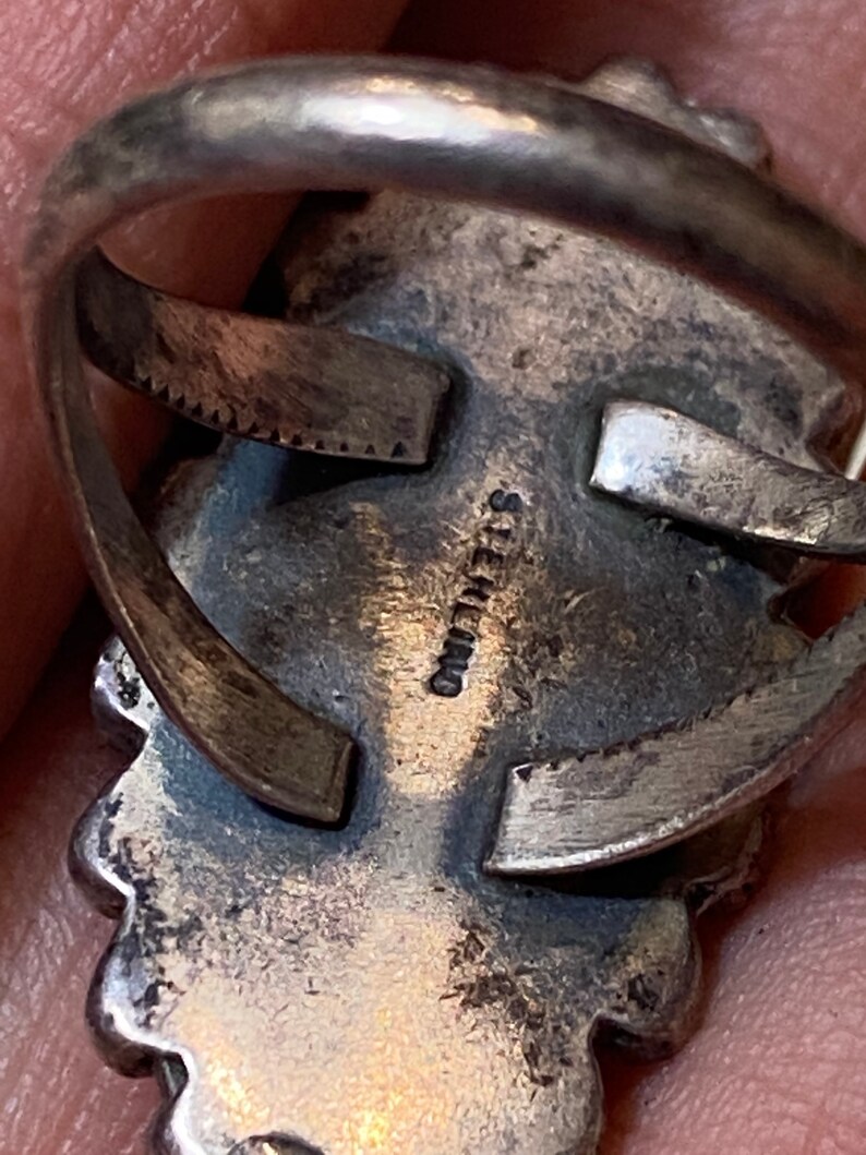 Puede incluir: Primer plano de la parte posterior de un anillo de plata con una inscripci&oacute;n estampada que dice "STERLING". El anillo tiene un dise&ntilde;o detallado con un borde festoneado.