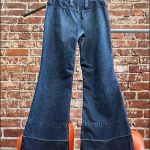 Pu&ograve; includere: Un paio di jeans a zampa di elefante in denim blu scuro con gamba larga e un bordo a contrasto blu scuro nella parte inferiore delle gambe.