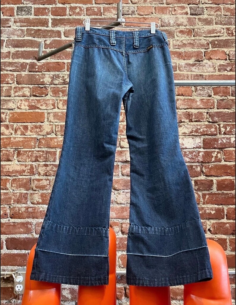 Pu&ograve; includere: Un paio di jeans a zampa di elefante in denim blu scuro con gamba larga e un bordo a contrasto blu scuro nella parte inferiore delle gambe.