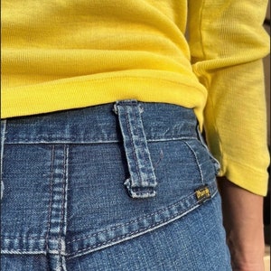 Pu&ograve; includere: Un paio di jeans blu denim con una camicia gialla infilata dentro. I jeans hanno una tasca posteriore con il logo Wrangler.